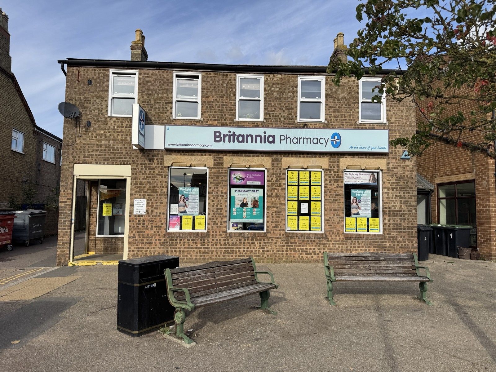 Britannia Pharmacy at 5 Market Square, Sandy, a brick building with a pale blue sign, flu and Covid-19 vaccination posters in the windows and two benches outside