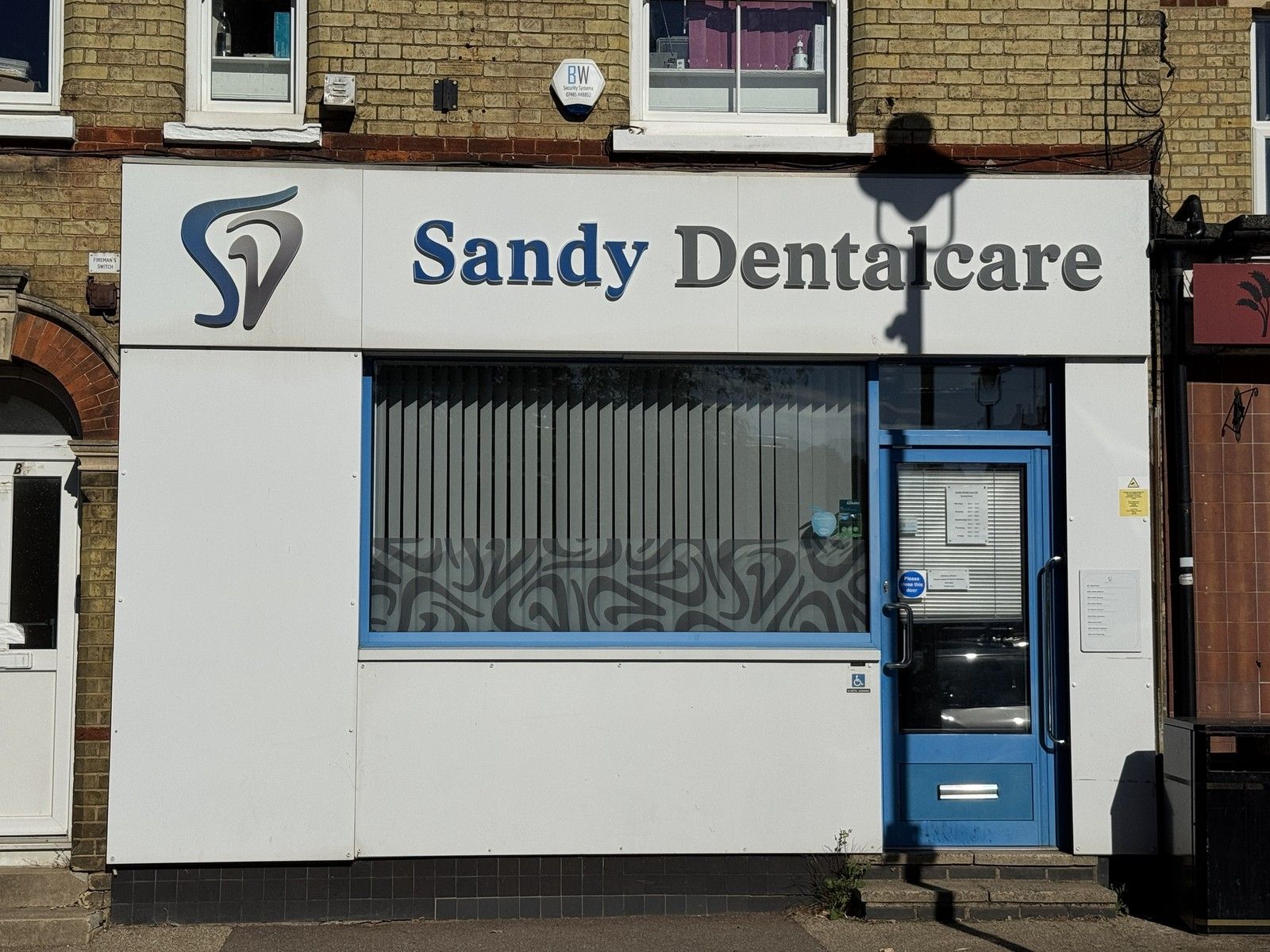 The shopfront of Sandy Dentalcare on Market Square, Sandy, with its white sign, blue and grey logo and blue front door